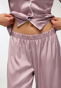 Set di pigiama in satin di un leggero color mauve, composto da una camicia con bottoni e rifiniture bianche e pantaloni con vita elastica. Texture liscia e vestibilità ampia.
