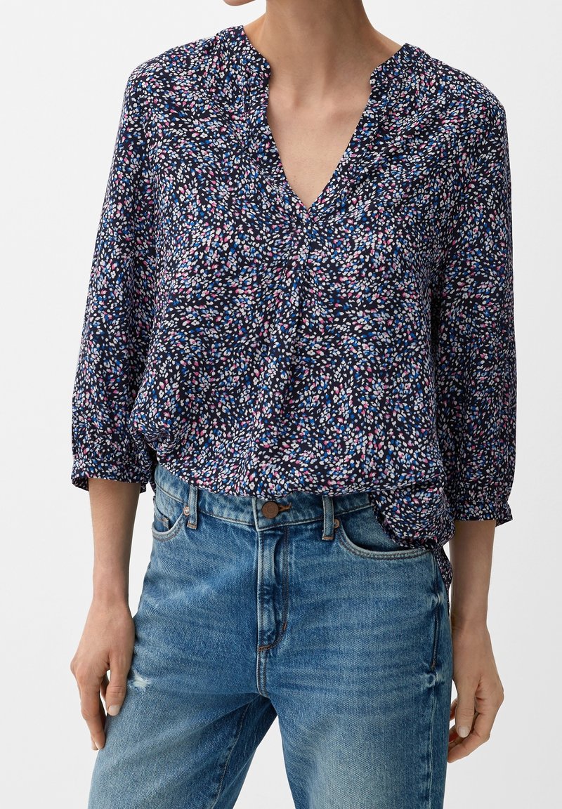 Femme portant un chemisier bleu marine à manches longues avec un petit motif floral multicolore et un jean bleu légèrement usé.