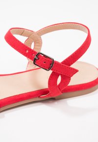 Sandale en daim rouge avec une semelle plate, dotée de brides croisées et d'une boucle en métal noir pour fixer la sangle à la cheville. Semelle intérieure beige clair.