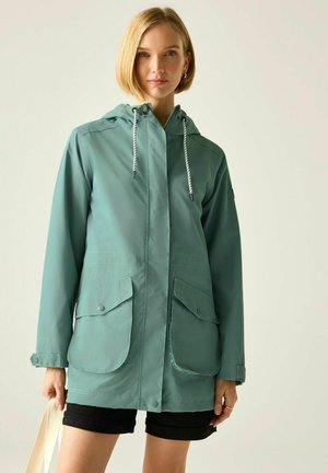Mujer que lleva un impermeable con capucha verde salvia con bolsillos delanteros y pantalones cortos negros, sosteniendo una bolsa beige contra un fondo liso.