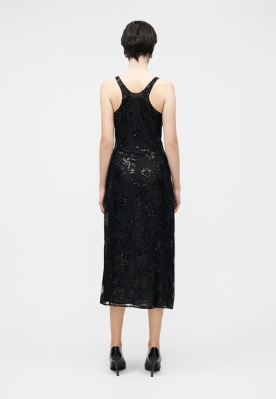 Robe noire sans manches avec un corsage ajusté et une jupe fluide, ornée de motifs d'étoiles en sequins et d'une superposition translucide. Texture lisse, longueur cheville.