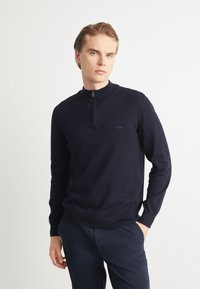 Marinblå zip-neck tröja i mjukt material, med en figursydd design och ribbade muddar. Diskret logotypdetalj på bröstet, tillsammans med marinblå byxor.