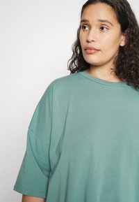 T-shirt oversized em algodão numa tonalidade de teal suave, com um decote redondo e mangas caídas, apresentando uma textura suave e costuras mínimas.