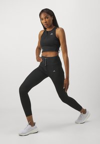Black athletic crop top with a front zipper. High-waisted black leggings featuring a decorative lace-up detail and logo accents. White sneakers.