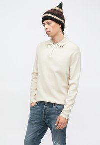 Πουλόβερ κρεμ με γιακά και φερμουάρ μέχρι τη μέση, κατασκευασμένο από μαλακό υλικό. Φορεμένο με τζιν με σκισίματα σε μπλε χρώμα και μοτίβο σκούφο.