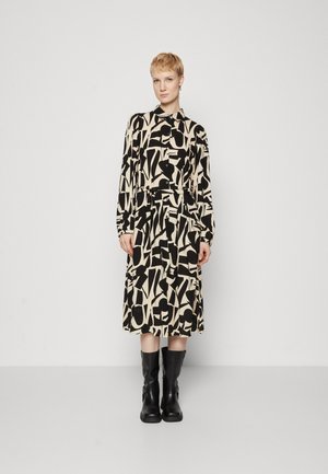 Long-sleeve dress in beige with a black abstract pattern, button closure, and pleated skirt. Paired with black knee-high boots.