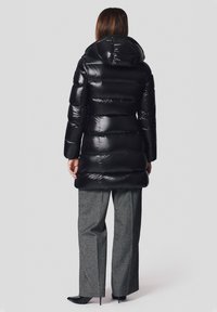 Czarna kurtka puchowa o błyszczącej powierzchni, pikowany wzór, odpinany kaptur i długość do bioder, w połączeniu z szarymi spodniami o szerokich nogawkach.