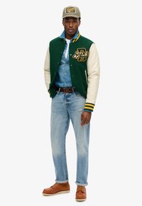 Chaqueta varsity verde y crema con puños a rayas amarillas, llevada sobre una camisa de mezclilla clara y jeans azules. Unos zapatos marrones con cordones completan el atuendo.