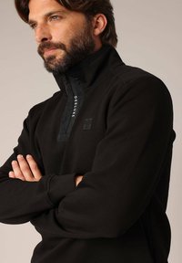 Zwarte fleece pullover met een halvemaanrits kraag, voorzien van een textuurpaneel aan de voorkant, logo-details en een aansluitend ontwerp.