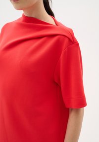 Röd topp med kort ärm som har en draperad krage, slät textur, avslappnad design och subtila sömnadsdetaljer längs ärmarna.