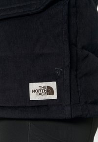 Gros plan sur l'ourlet d'une veste en velours côtelé noire avec un patch logo The North Face blanc et marron cousu près de la couture.