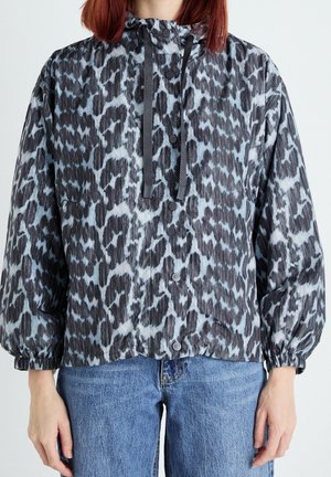 Frau trägt eine schwarz-grau gemusterte Kapuzenjacke mit elastischen Bündchen und blaue Jeans, steht mit entspannten Armen seitlich am Körper.