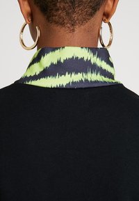 Primer plano del cuello y oídos de una persona, que lleva grandes pendientes de aro de oro y una blusa negra con un cuello de patrón verde y negro.