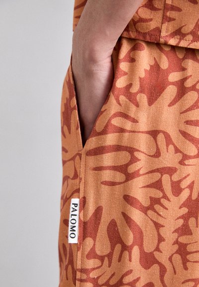 Tissu orange avec des formes organiques abstraites en orange plus foncé. Poche sur le côté avec une étiquette blanche affichant "PALOMO." Texture lisse.