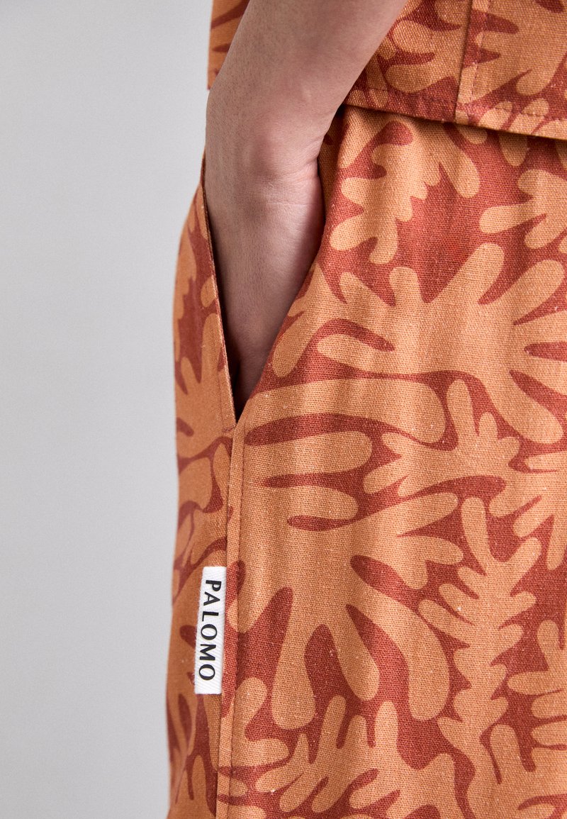 Orange fabric featuring abstract organic shapes in darker orange. Side seam pocket with a white label displaying "PALOMO." Smooth texture.