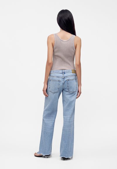 Femme aux longs cheveux noirs portant un top beige sans manches, un jean large bleu clair avec des ourlets effilochés et des sandales noires, debout de dos.