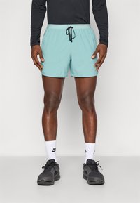 Turquoise sportshorts met een elastische tailleband en een trekkoord, met twee zijzakken. Gecombineerd met zwarte hardloopschoenen en witte sokken.