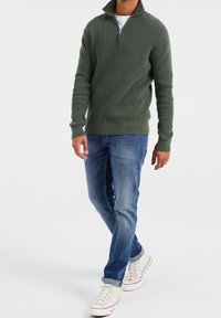 Maglione a coste verde con zip a quarti, abbinato a jeans azzurri chiari e sneakers bianche alte. Tessuto morbido con uno stile aderente.