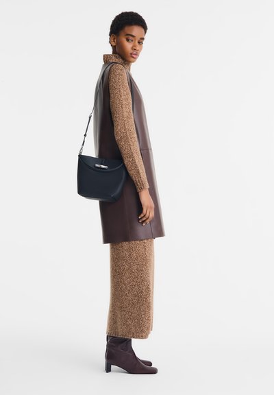 Braunes Lederkleid ohne Ärmel über einem strukturierten braunen Strickrock, kombiniert mit einer schwarzen Tasche und braunen Absatzstiefeln.