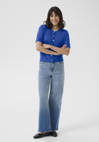 Cardigan bleu à manches courtes avec fermeture à boutons, associé à un jean large délavé et des ballerines noires, présentant une coupe décontractée et une texture lisse.