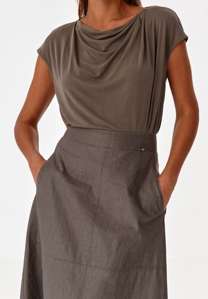 Femme portant un haut court drapé taupe rentrée dans une jupe grise taille haute, les mains dans les poches.