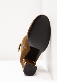 Bottine en suede marron avec semelle en caoutchouc noir, fermeture éclair dorée et talon bloc décoratif avec accents dorés, positionnée debout contre des murs blancs.