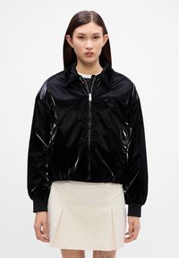 Jachetă bomber neagră, lucioasă, cu fermoar în față, gulere oversized, manșete elastice și croială scurtă. Pur tată peste o fustă crem.