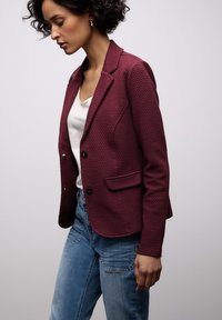 Blazer texturé marron rouge avec une coupe ajustée, doté d'un col cranté, de deux poches avant et de boutons noirs ; associé à un jean en denim bleu.
