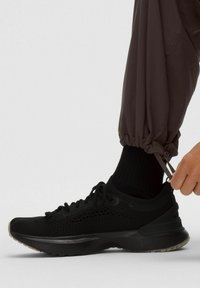 Pantofii sport negri, cu un superior din plasă și talpă din cauciuc, sunt asortați cu o pereche de pantaloni sport din material închis. O mână ajustează șireturile din lateral.