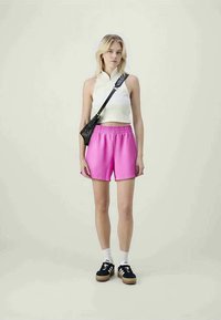 Shorts roses avec une taille élastique, associés à un haut sans manches blanc. Sac en bandoulière noir, chaussettes blanches au niveau des chevilles et baskets noires.