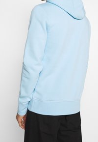 Světle modrá mikina s kapucí se šňůrkami, žebrovanými manžetami a lemem, vyrobená z měkkého materiálu. Má volný střih a jednoduchý design.