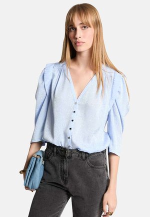 Blusa azzurro chiaro con scollo a V, maniche a sbuffo e dettagli con bottoni, abbinata a jeans in denim grigio scuro e una piccola borsa blu.