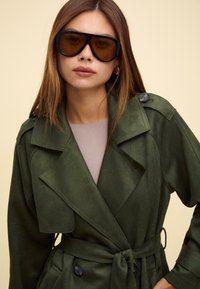 Maslinasto zelena faux suede trench kaput s velikim reverima, remenom za vezanje i dugmadi kao detaljima. Nosite ga s velikim tamnim naočalama.