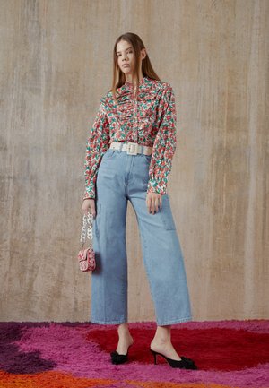 Camisa de manga longa com padrão floral, jeans de perna larga azul claro, cinto branco, mules pretos de salto, e pequena bolsa vermelha com padrão. Fundo brilhante.