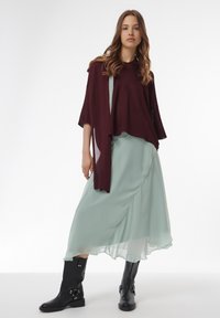 Bordeaux gebreide trui, losvallend, gedragen over een vloeiende mintgroene maxi-jurk. Zwarte laarzen tot halverwege de kuit met gespen en studs.