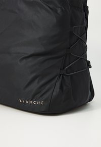 Juodas nailono maišelis su susiraukšlėjusiomis šonų detalėmis, reguliuojamomis virvėmis ir šviesiu "BLANCHE" logotipu apačioje.