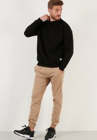 Zwarte sweatshirt met ronde hals, beige joggerbroeken en zwarte sportschoenen. Casual outfit met elastische boorden en een klein logo op de sweatshirt.
