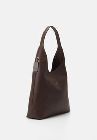 Sac à bandoulière en cuir marron foncé avec large bandoulière, poche avant avec petit bouton doré et étiquette attachée sur fond uni.