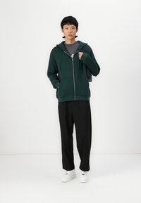 Un hoodie zippé vert avec des cordons de serrage, superposé à un t-shirt gris, associé à un pantalon noir et des baskets blanches, se tenant devant un fond blanc.