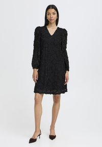 Svart klänning med strukturerat diamantmönster, djup V-ringning, långa ärmar och en flödande design. Matchad med mörka spetsiga klackskor.