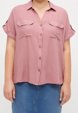 Person wearing a loose dusty pink short-sleeve button-up shirt with flap chest pockets and rolled sleeves, paired with a blue denim skirt.