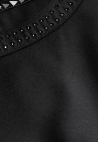 Zwarte stoffen top met een gestructureerde halslijn voorzien van kleine zwarte studs en een contrasterende patronen binnenafwerking. Gladde textuur, strakke design.