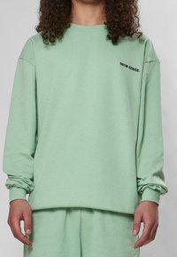 Felpa in cotone verde menta con maniche lunghe, scollo rotondo e dettaglio del logo ricamato sul petto. Tessuto morbido, vestibilità comoda.