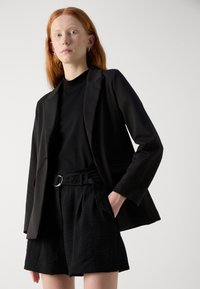 Black tailored blazer over a black turtleneck, paired with loose black shorts featuring a buckle belt. Smooth fabric with a structured design.