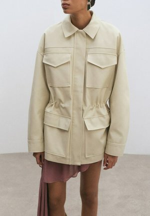 Femme portant une veste beige oversize avec quatre grandes poches à rabat et une taille cintrée, associée à une jupe asymétrique mauve.