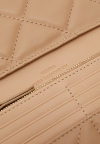 Beige prešívaná koža s kovovým zipsom v zlatej farbe a prešitím. Zobrazuje embosovanú značku a logo. Hladká textúra s lineárnymi akcentmi.