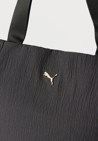 Sac en tissu texturé noir avec un logo Puma doré et de larges poignées noires sur un fond uni.