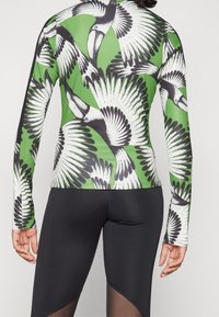 Långärmad topp med grön botten och tryck av svart-vita tukaner. Matchas med svarta leggings med texturerad design och utskärningar.