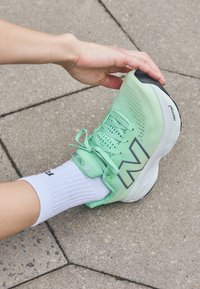 Light green running shoe with breathable mesh upper, grey accents, and black sole. Worn with a white, ribbed sock against a stone surface.