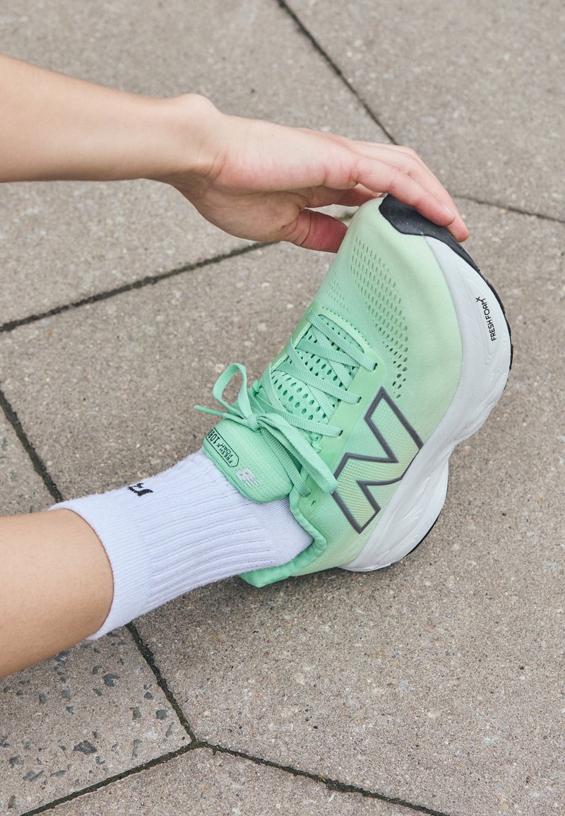 Light green running shoe with breathable mesh upper, grey accents, and black sole. Worn with a white, ribbed sock against a stone surface.